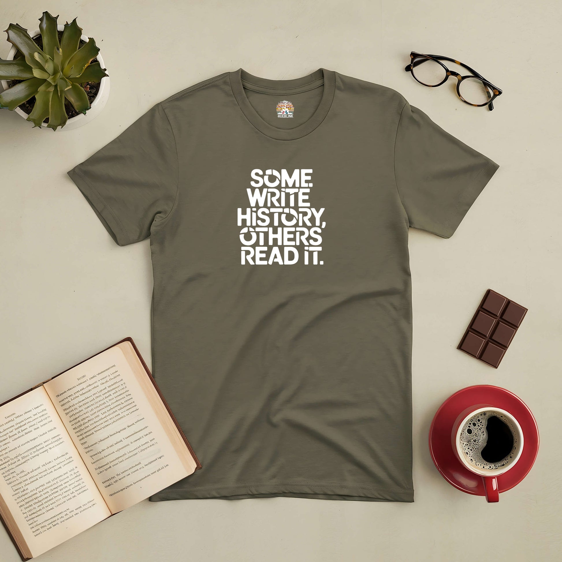 Green t-shirt with "Some Write History, Others Read It" text, surrounded by an open book, glasses, chocolate, and a coffee cup.