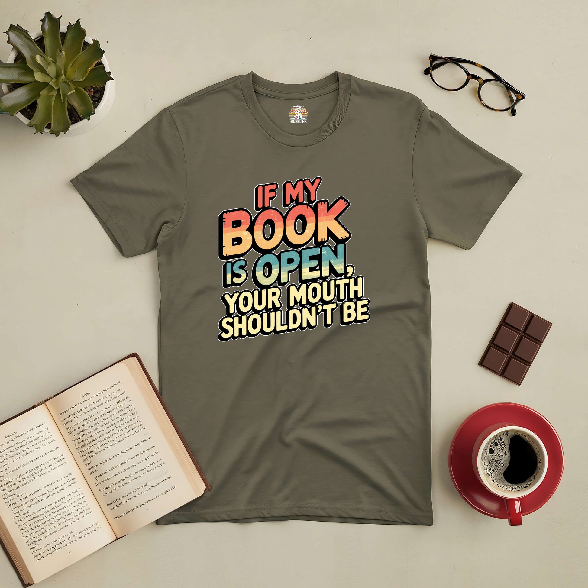 Olive green tee with text "If My Book Is Open, Your Mouth Shouldn't Be," surrounded by a book, coffee, glasses, and chocolate.