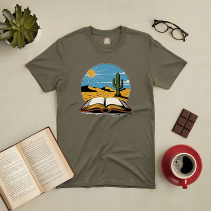 Silent Desert Tee with cactus and open book design, surrounded by coffee, chocolate, glasses, and a potted plant, on a table.