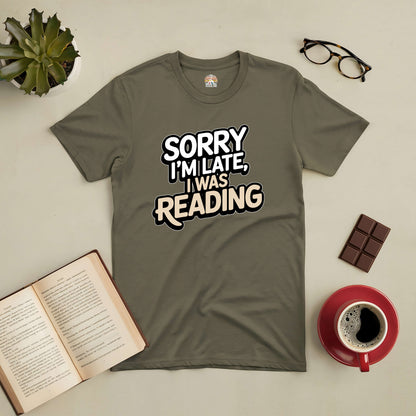 Olive green "Sorry I'm Late, I Was Reading" t-shirt displayed with a book, coffee, eyeglasses, chocolate, and a plant.