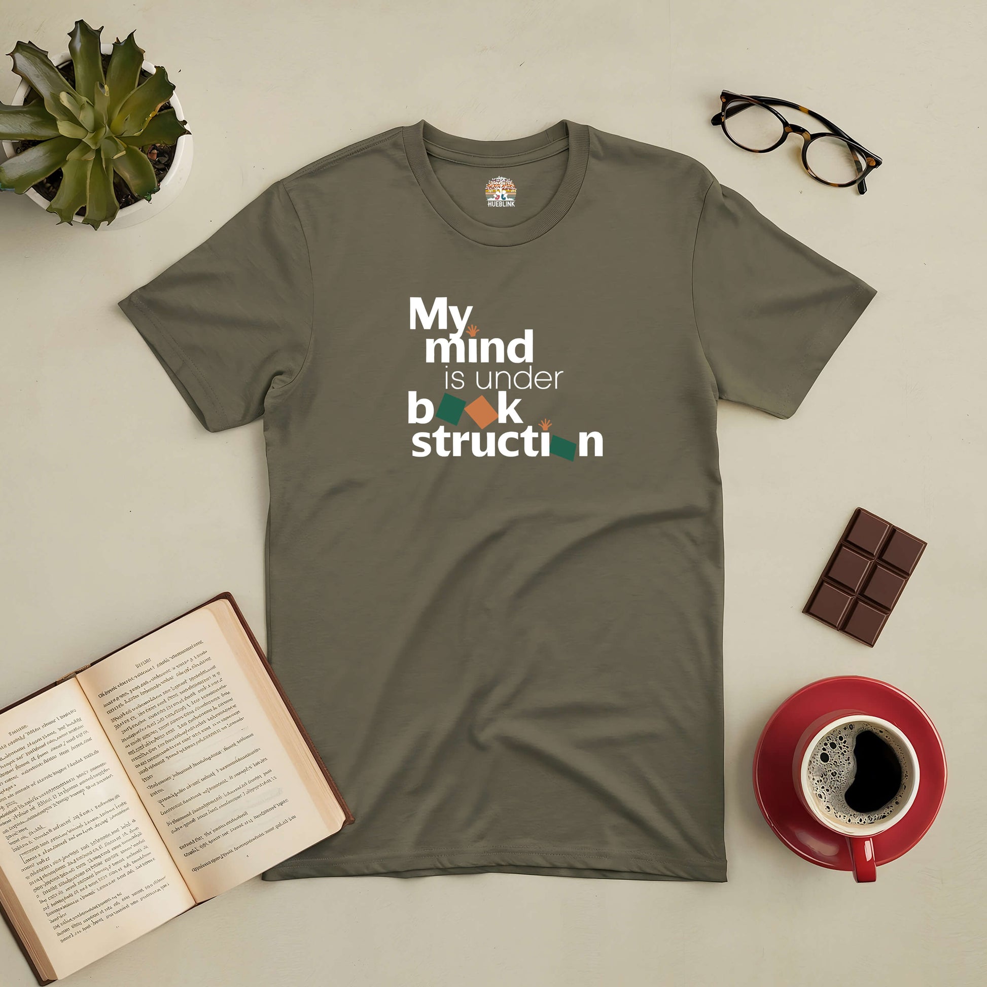 Olive green tee with "My Mind is Under Book-struction" design, surrounded by an open book, glasses, chocolate, and a coffee cup.