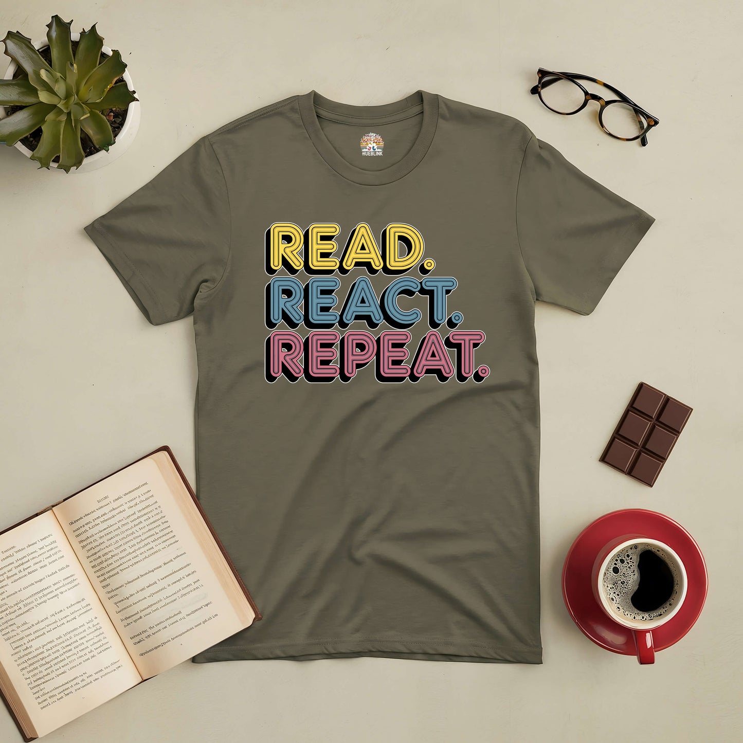 "Read React Repeat Tee with colorful text, surrounded by coffee, open book, glasses, and chocolate on a neutral background"