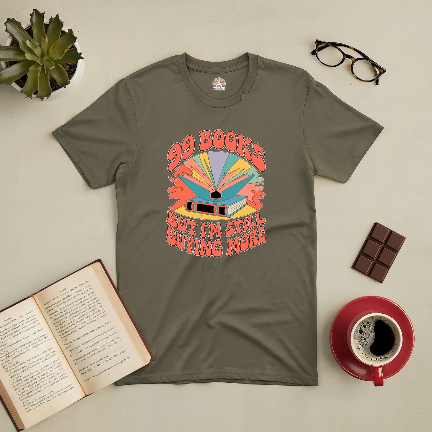 "99 Books and Counting! Tee with colorful book graphic design, perfect for book lovers. Displayed with open book, coffee, and chocolate."