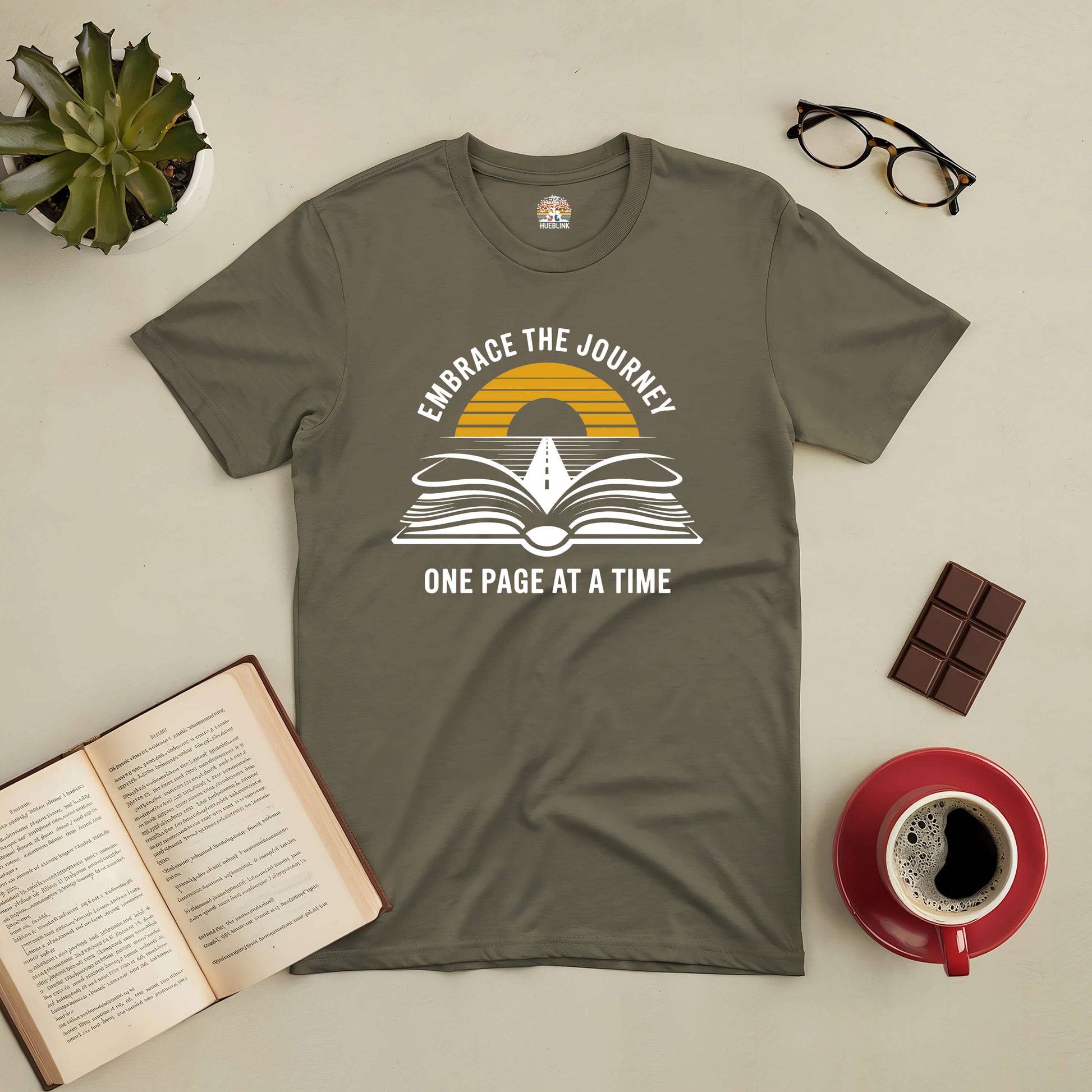 Embrace the Journey t-shirt with book design, road to horizon, surrounded by coffee, book, glasses, chocolate, and plant.