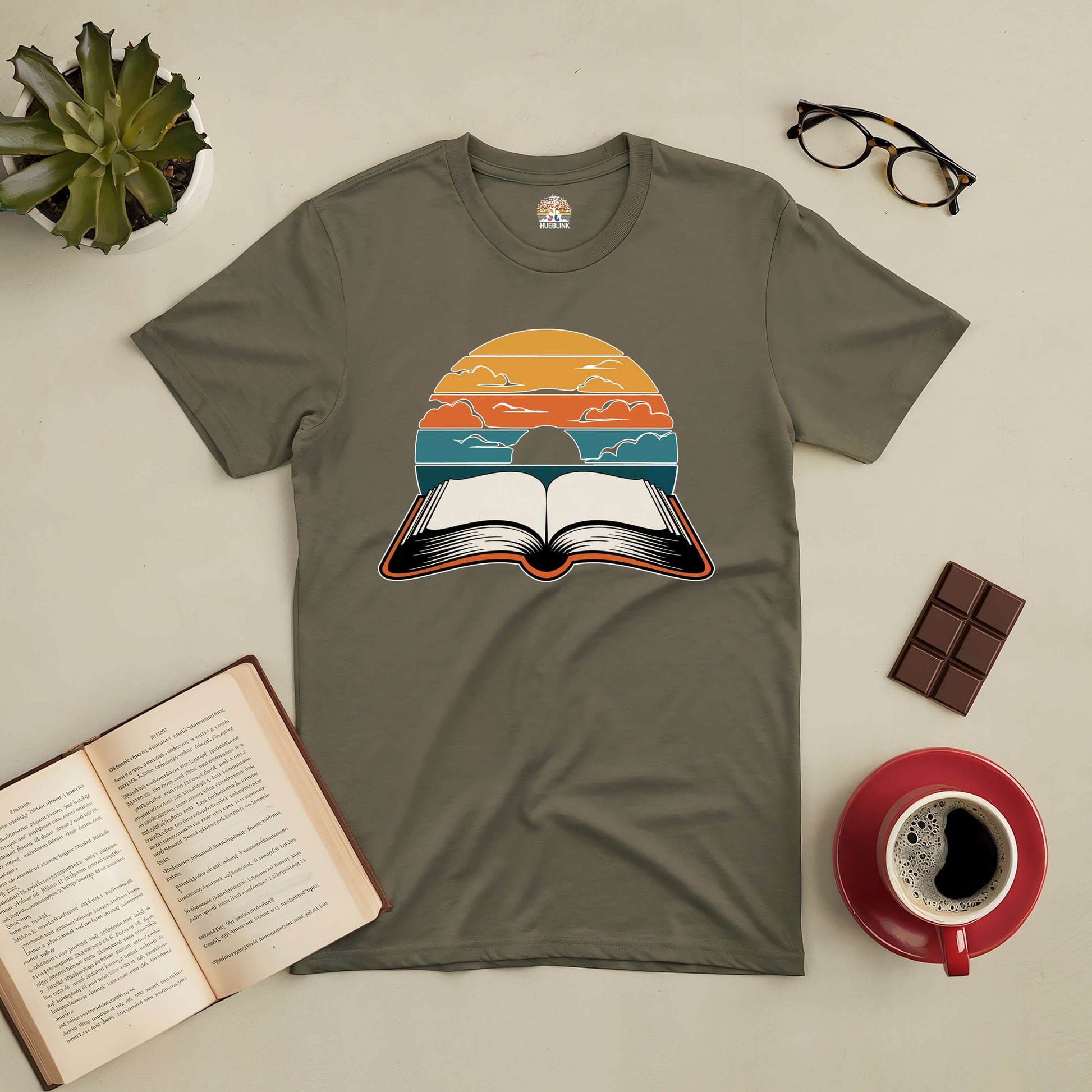 Olive green tee featuring an open book under a vibrant, colorful sky, symbolizing the imaginative worlds of reading.