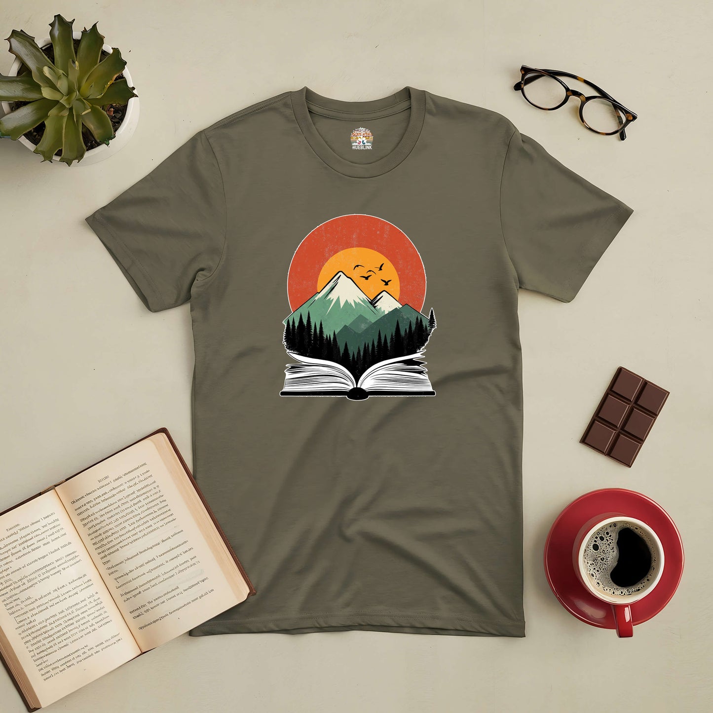 Green t-shirt with mountain and book design, surrounded by a plant, open book, glasses, chocolate, and coffee cup.