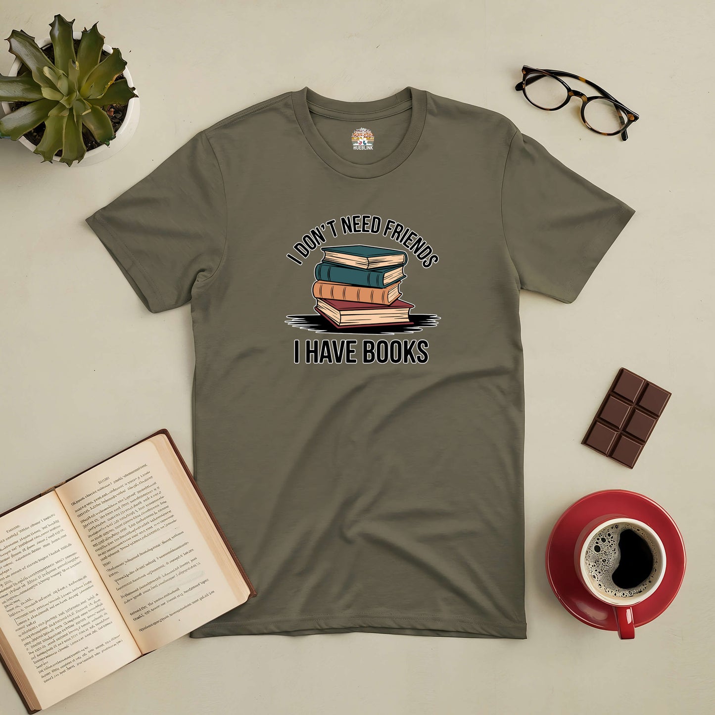 Olive green tee with "I Don't Need Friends, I Have Books" text and book graphic, surrounded by coffee, chocolate, glasses, and an open book.