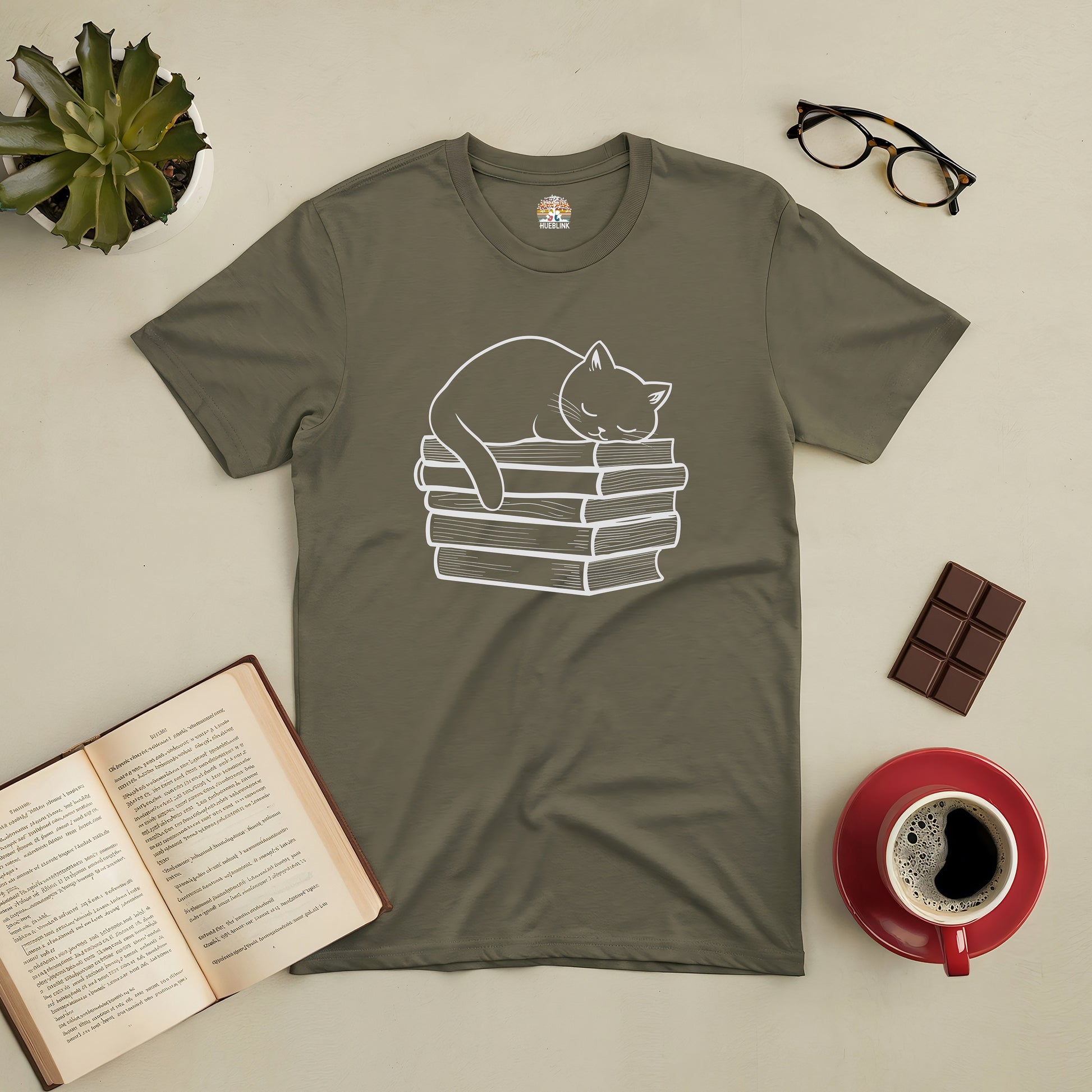 Olive green tee featuring a graphic of a cat napping on a stack of books, surrounded by coffee, chocolate, and glasses for cozy vibes.