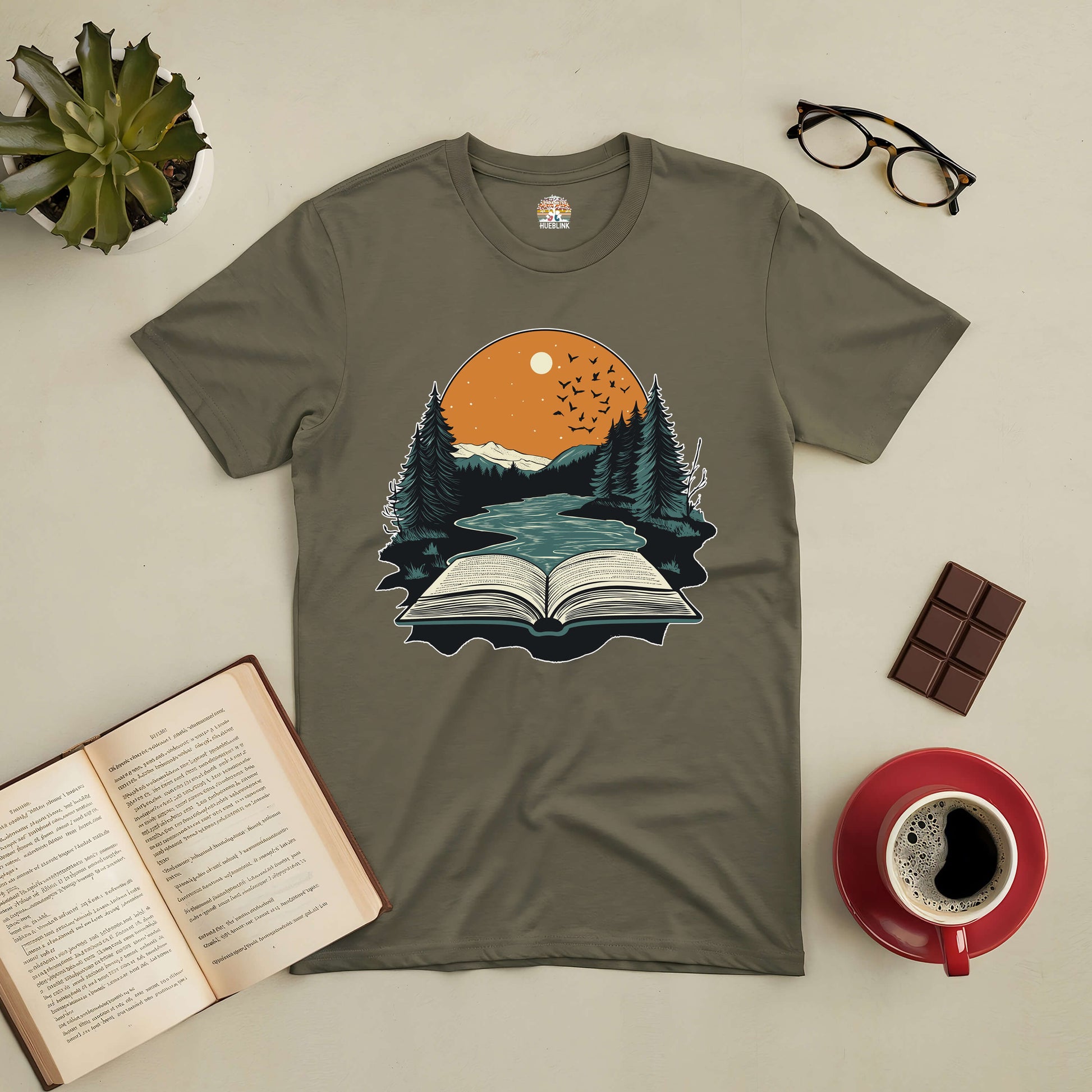 Absorbed in Nature tee featuring an open book and forest landscape design, surrounded by a coffee cup, glasses, and chocolate.