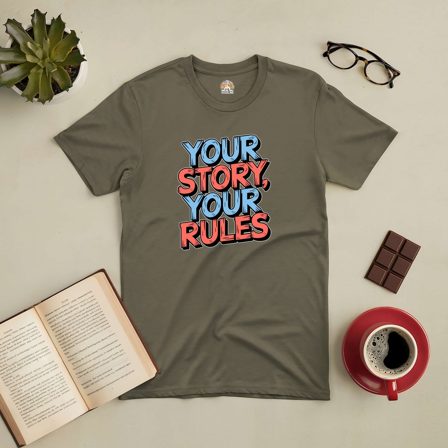 Sage green tee with "Your Story, Your Rules" text, surrounded by a book, glasses, chocolate, and coffee, promoting individuality.