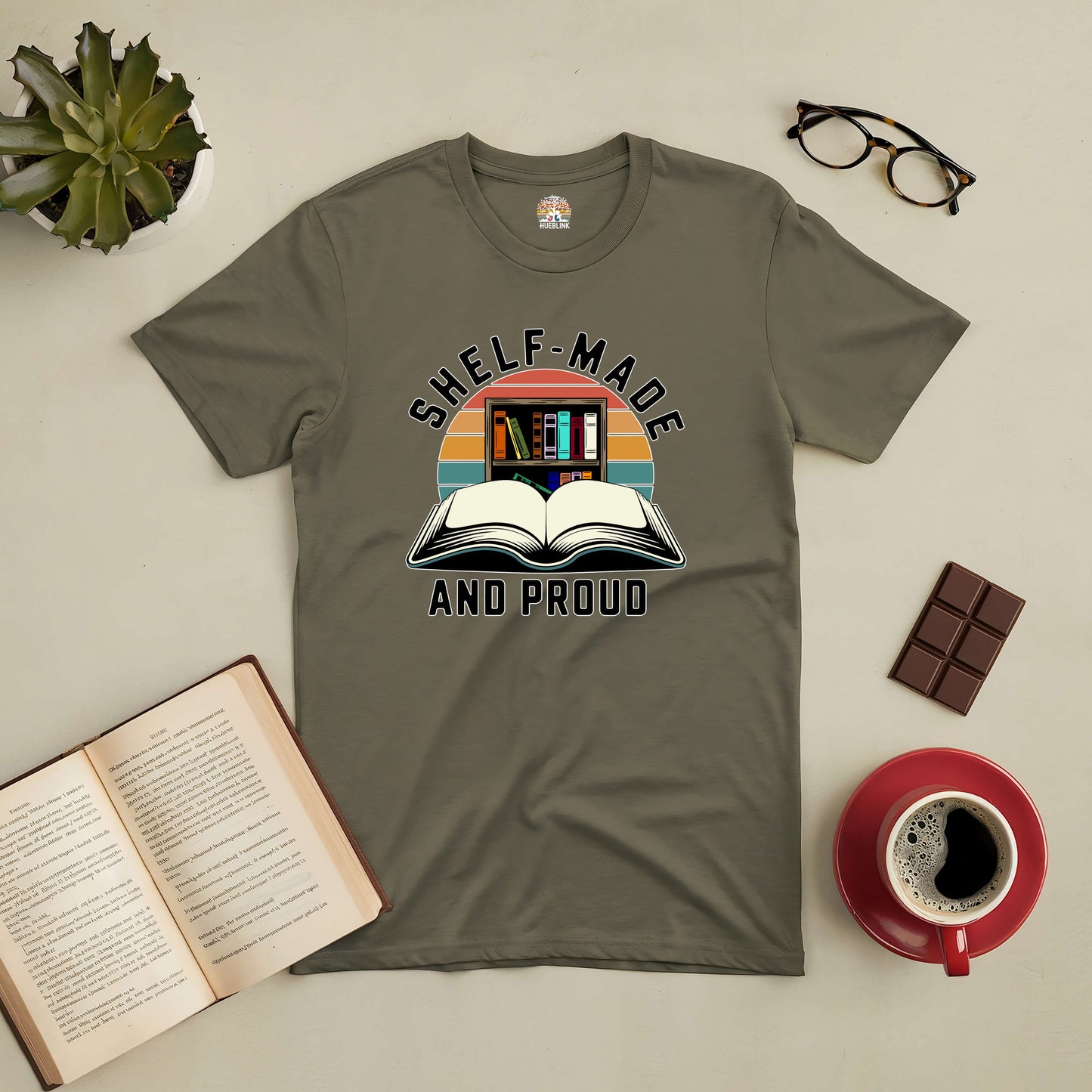 "Shelf-Made And Proud tee for book lovers with open book and bookshelves graphic, laid out with coffee, chocolate, and glasses."