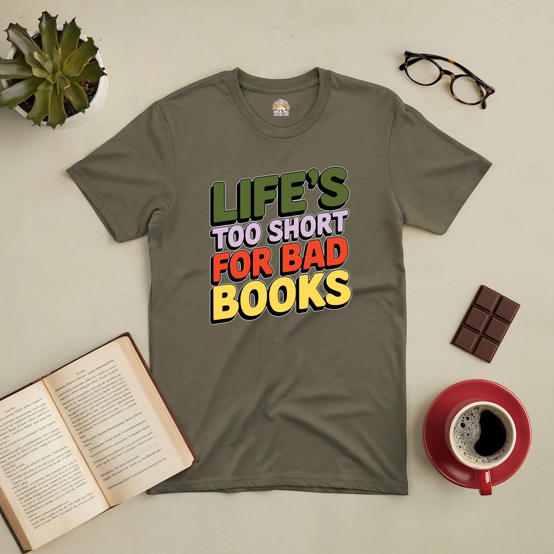 Olive green tee with "Life's Too Short for Bad Books" text, surrounded by a book, glasses, chocolate, plant, and a coffee cup.