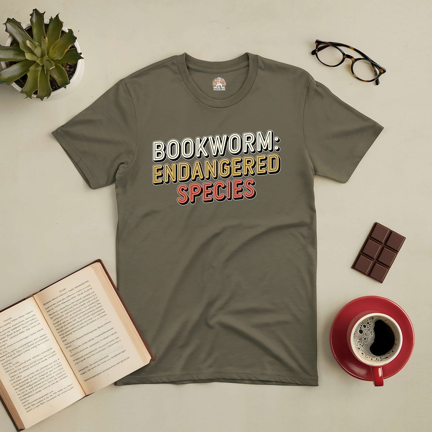 Green t-shirt with "Bookworm: Endangered Species" text, surrounded by an open book, glasses, coffee cup, chocolate, and plant.