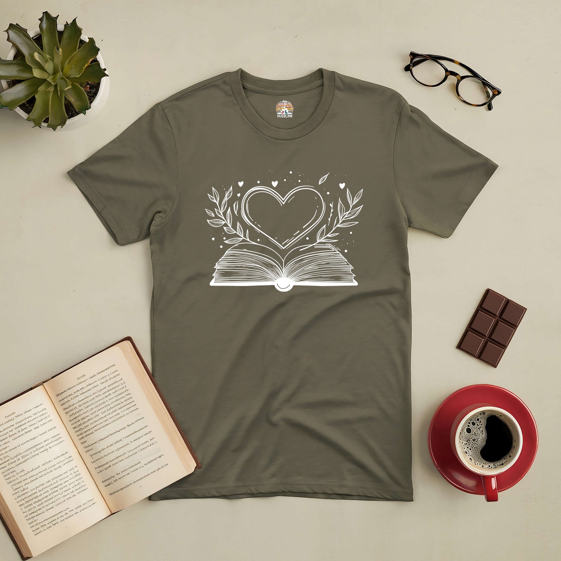 Love Stories Tee with heart and book design, surrounded by coffee, glasses, chocolate, open book, and plant. Perfect for book lovers.