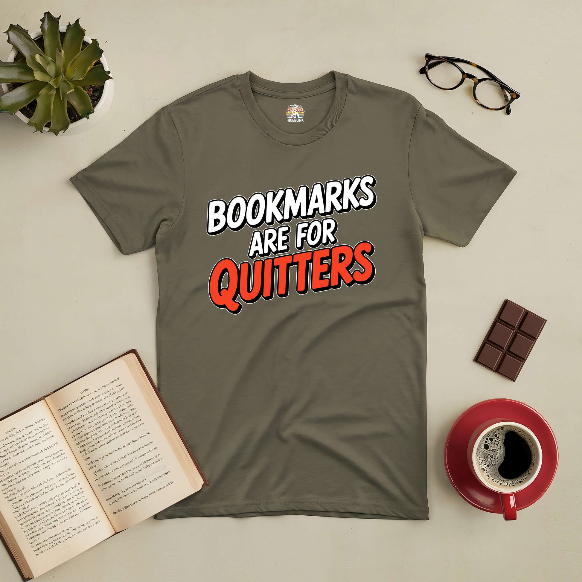 Olive green tee with bold text "Bookmarks Are for Quitters," surrounded by a book, glasses, chocolate, and coffee on a table.