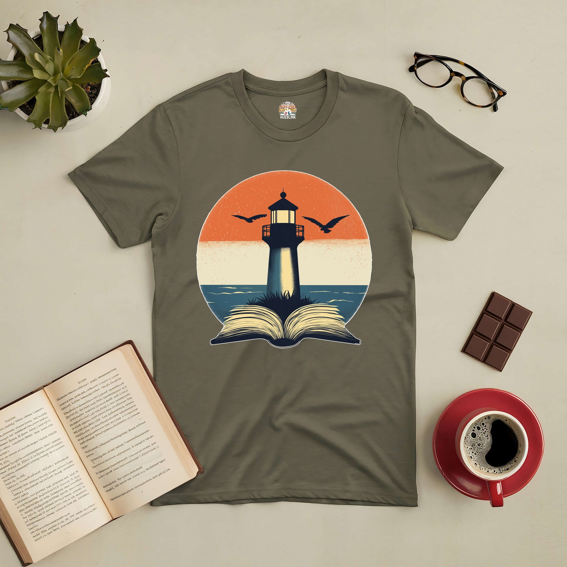 Olive green tee with a lighthouse illuminating an open book, symbolizing exploration and wisdom. Nearby are coffee, glasses, and chocolate.