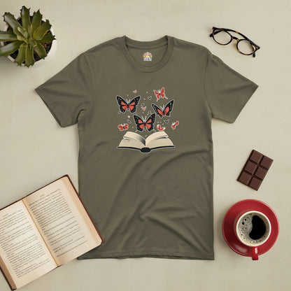 Green tee with open book and butterfly design, surrounded by glasses, chocolate, coffee, and a potted plant, themed "Where Stories Take Flight".