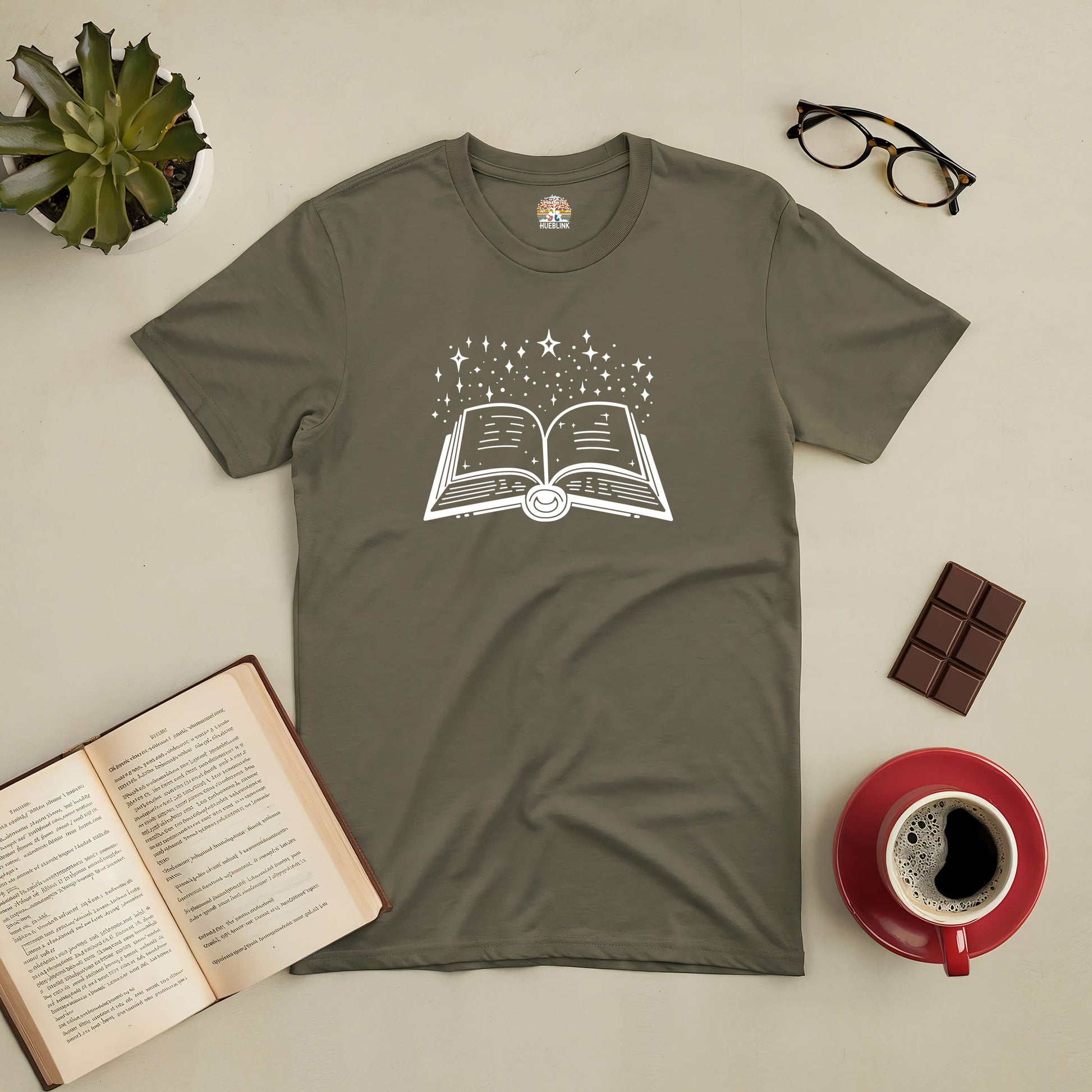 Olive green Stellar Script Tee with open book and star design, accompanied by glasses, chocolate, coffee, and an open book.