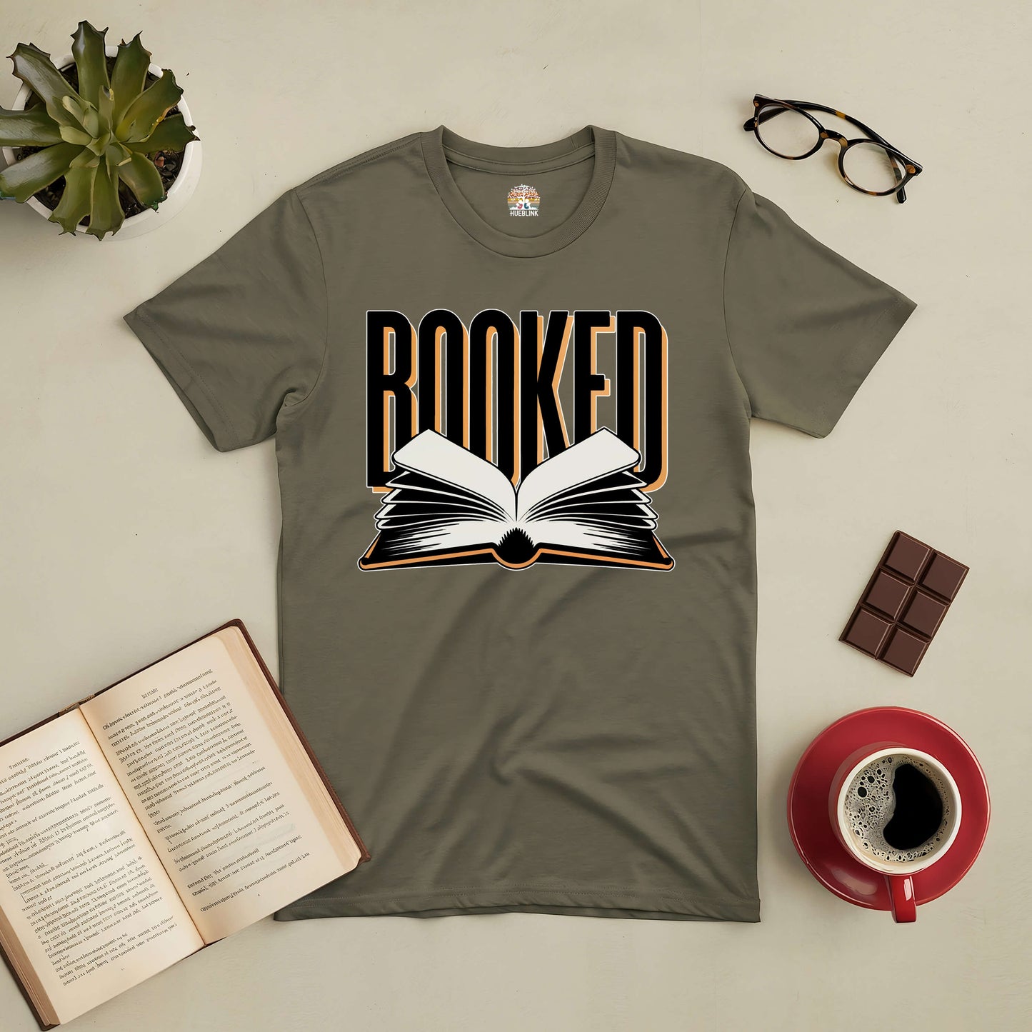 Olive green "Booked" tee with open book graphic laid flat, surrounded by an open book, glasses, a chocolate bar, and coffee cup.