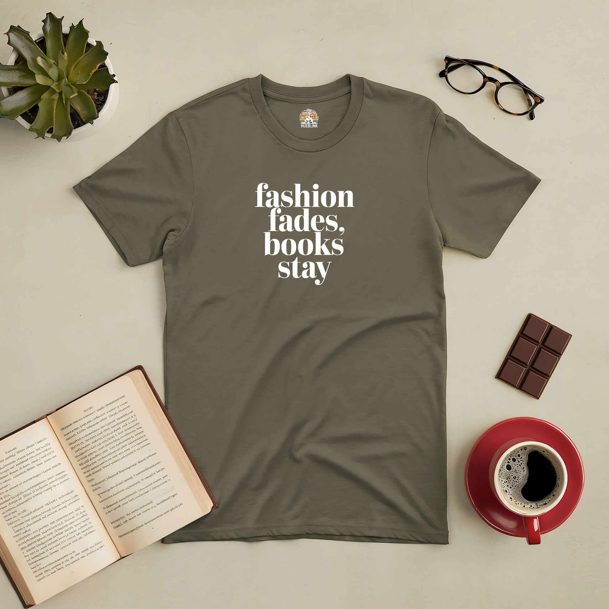 Olive green tee with "Fashion fades, books stay" print, styled with an open book, glasses, chocolate, and coffee.