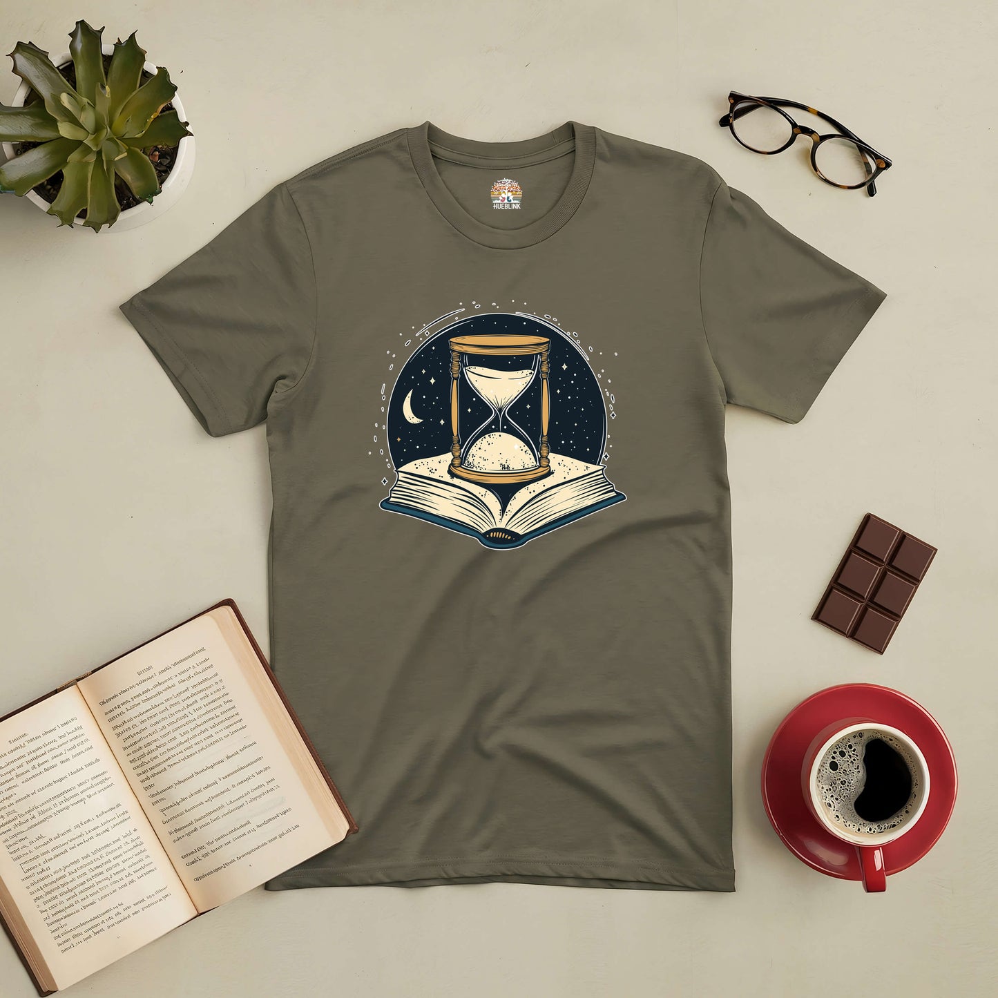 "Chronicles of Time and Space Tee with hourglass and book design, surrounded by coffee, chocolate, and a succulent plant."