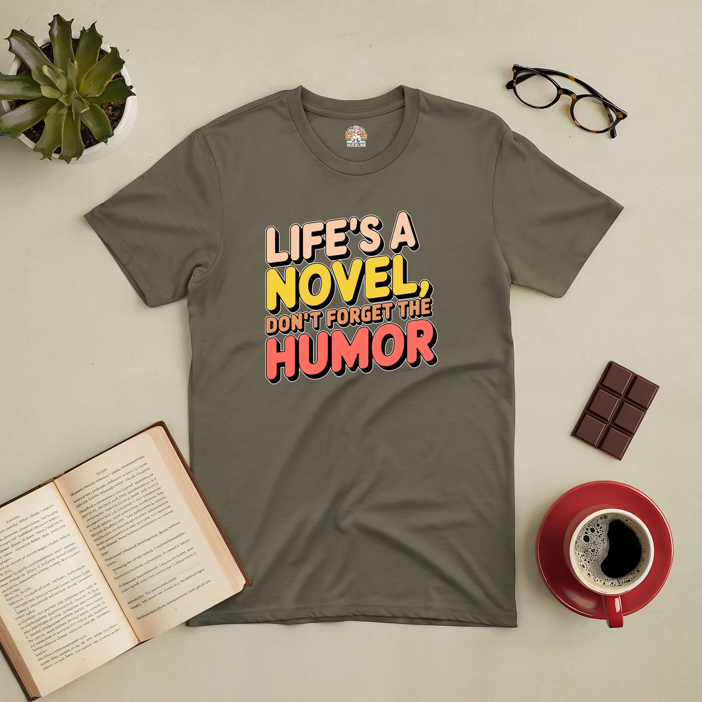 Grey tee with "Life's a Novel, Don't Forget the Humor" text, surrounded by glasses, book, chocolate, and coffee cup on a table.