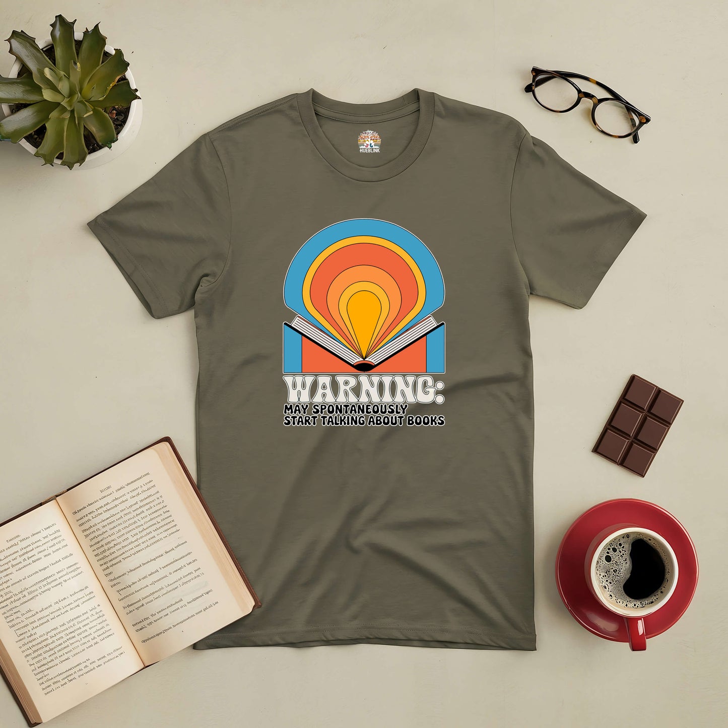 "Warning: May Start Talking About Books Tee with colorful book graphic, surrounded by open book, coffee, glasses, plant, and chocolate"