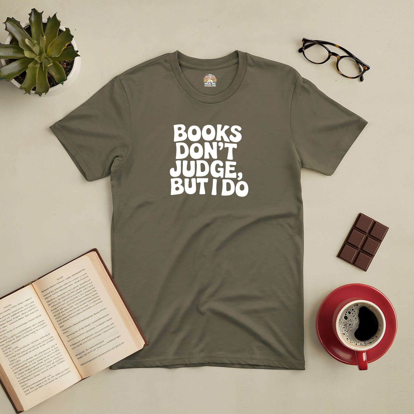 Olive green tee with "Books Don't Judge, But I Do" slogan, surrounded by an open book, glasses, chocolate, and a coffee cup.