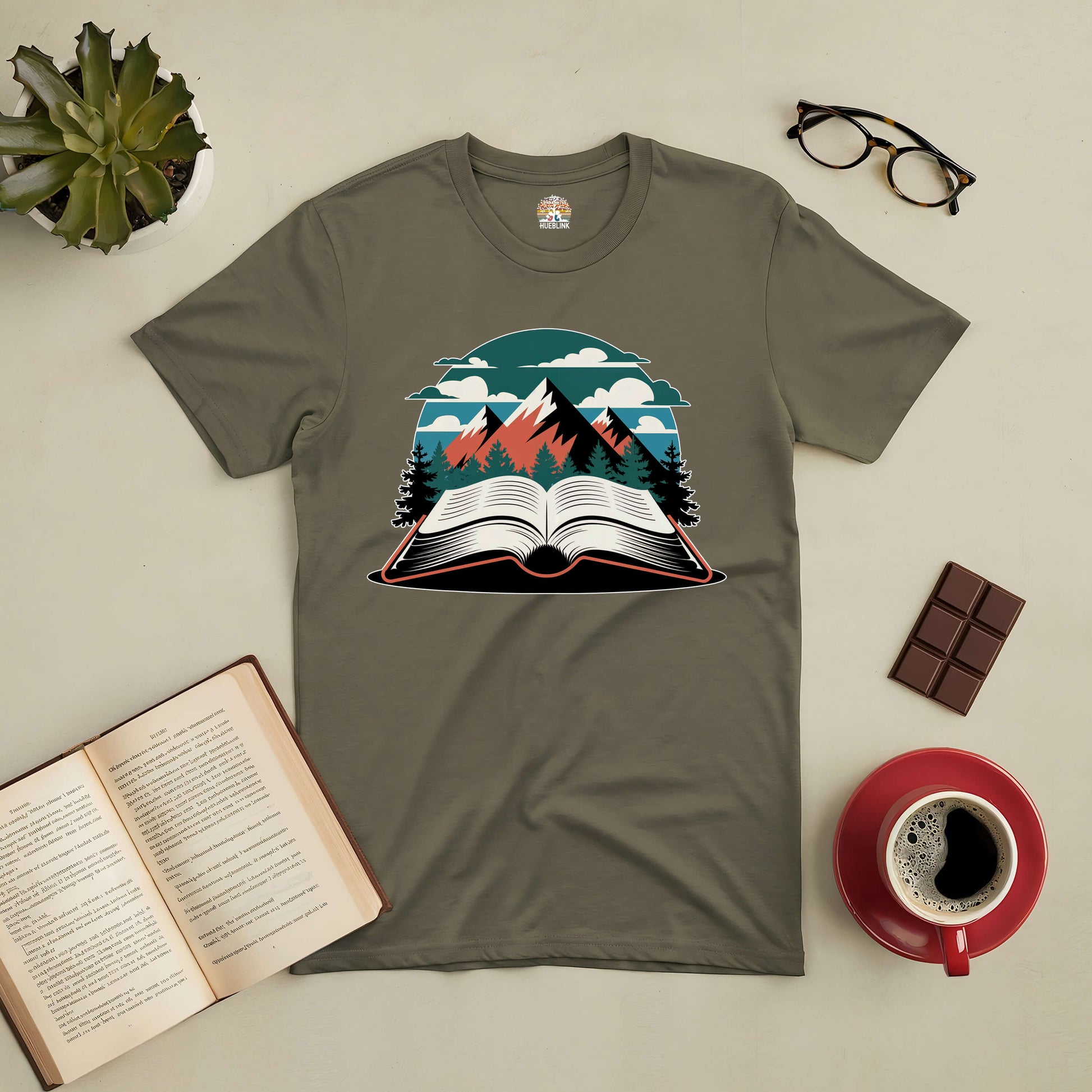 "Summits of Inspiration Tee with book and mountain design, surrounded by open book, coffee, glasses, chocolate, and plant"