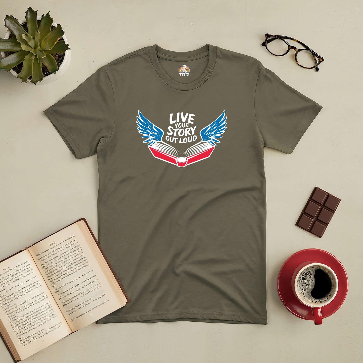 Olive green tee with open book and wings design, text "Live Your Story Out Loud," perfect for dreamers and storytellers, coffee and book nearby.