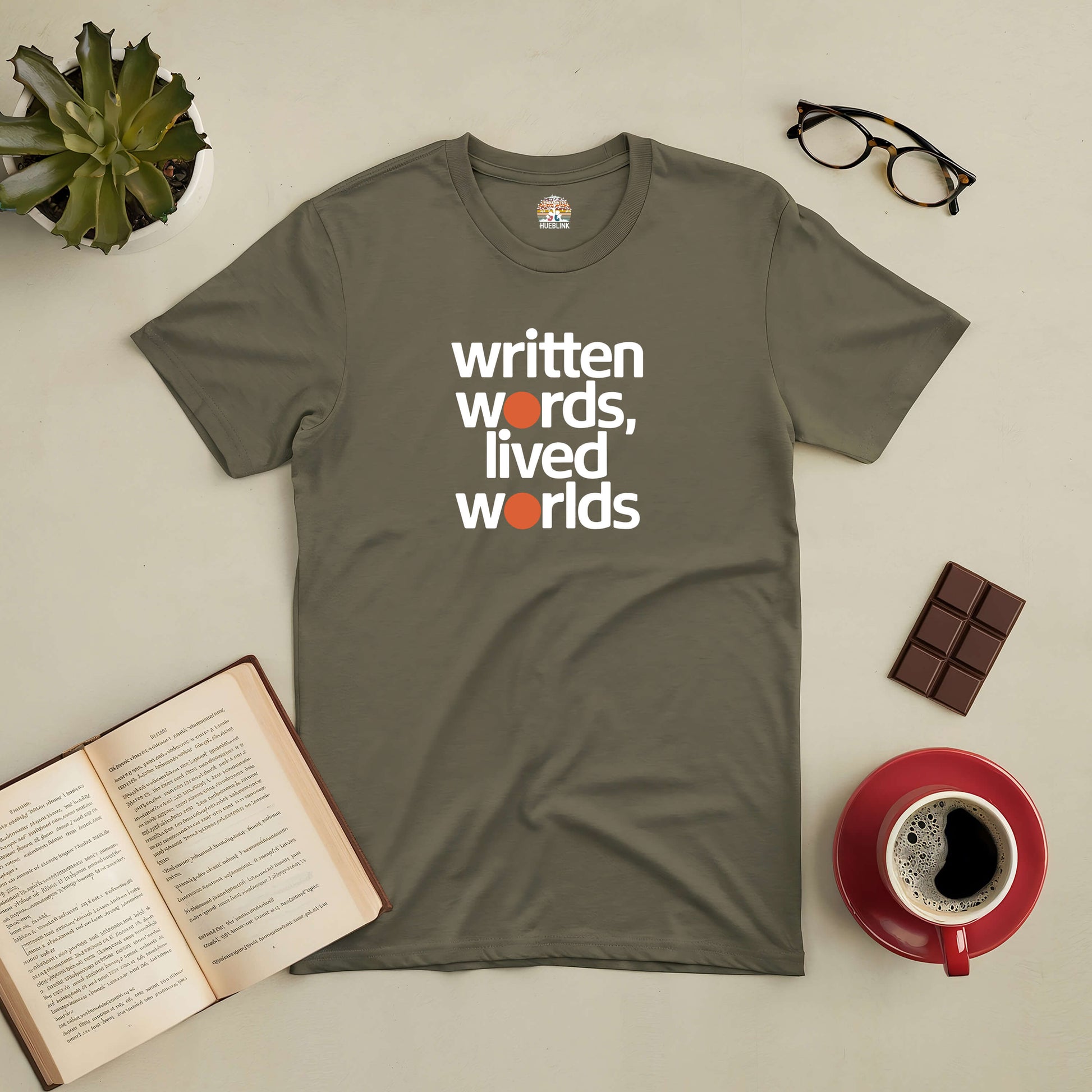 Olive green "Written Words, Lived Worlds" tee with coffee, chocolate, book, glasses, and a plant on a neutral background.