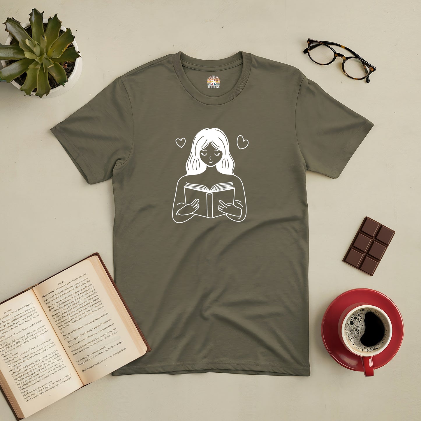 Bookish Bliss Tee with reading illustration, styled with an open book, coffee, and chocolate on a cozy flat lay.