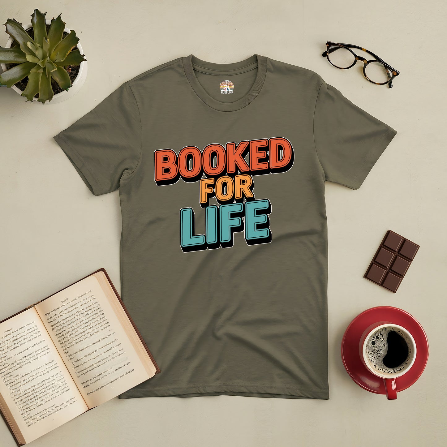 "Booked for Life Tee with colorful text, perfect for book lovers, surrounded by open book, glasses, coffee, and chocolate"