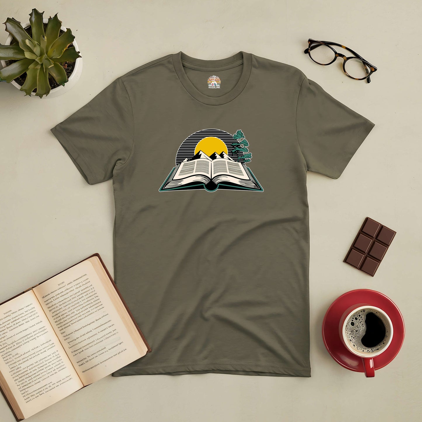 "Where Every Story Shines Tee with book and sunrise design, surrounded by coffee, chocolate, glasses, and an open book"