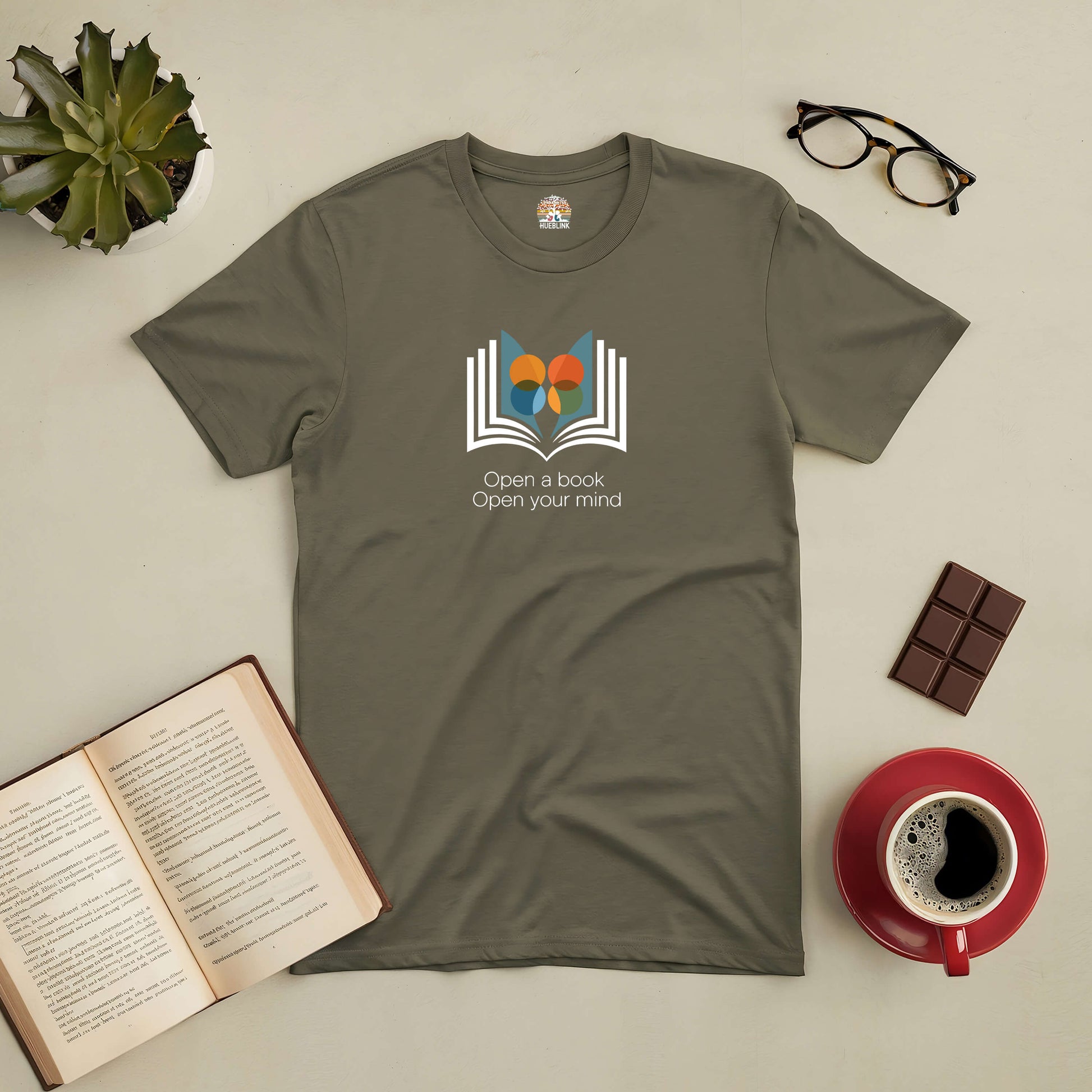 Olive green tee with "Open a Book, Open Your Mind" design, surrounded by a book, coffee, glasses, and a plant.