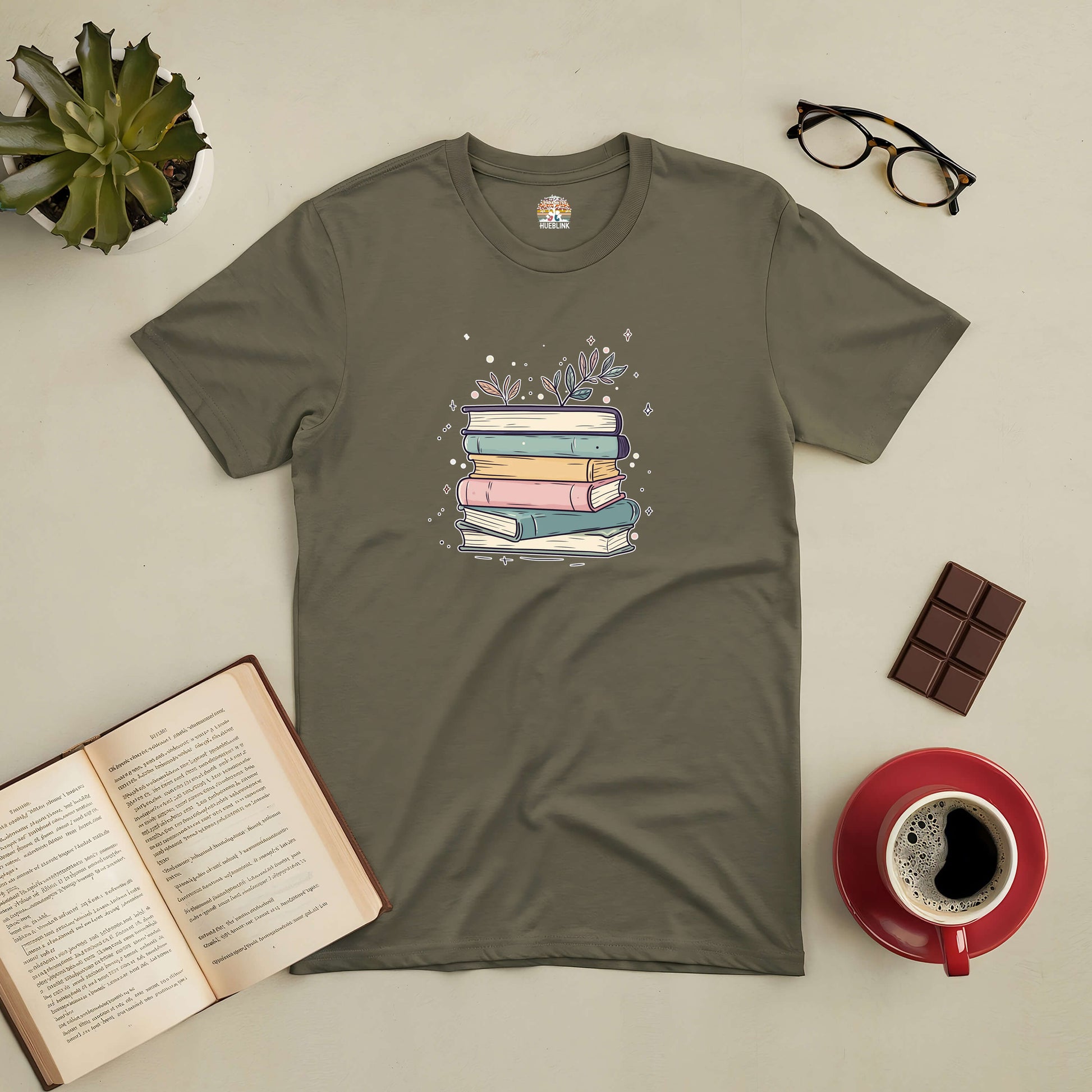 Olive green tee with pastel-colored books and floral accents, surrounded by an open book, coffee cup, and glasses.