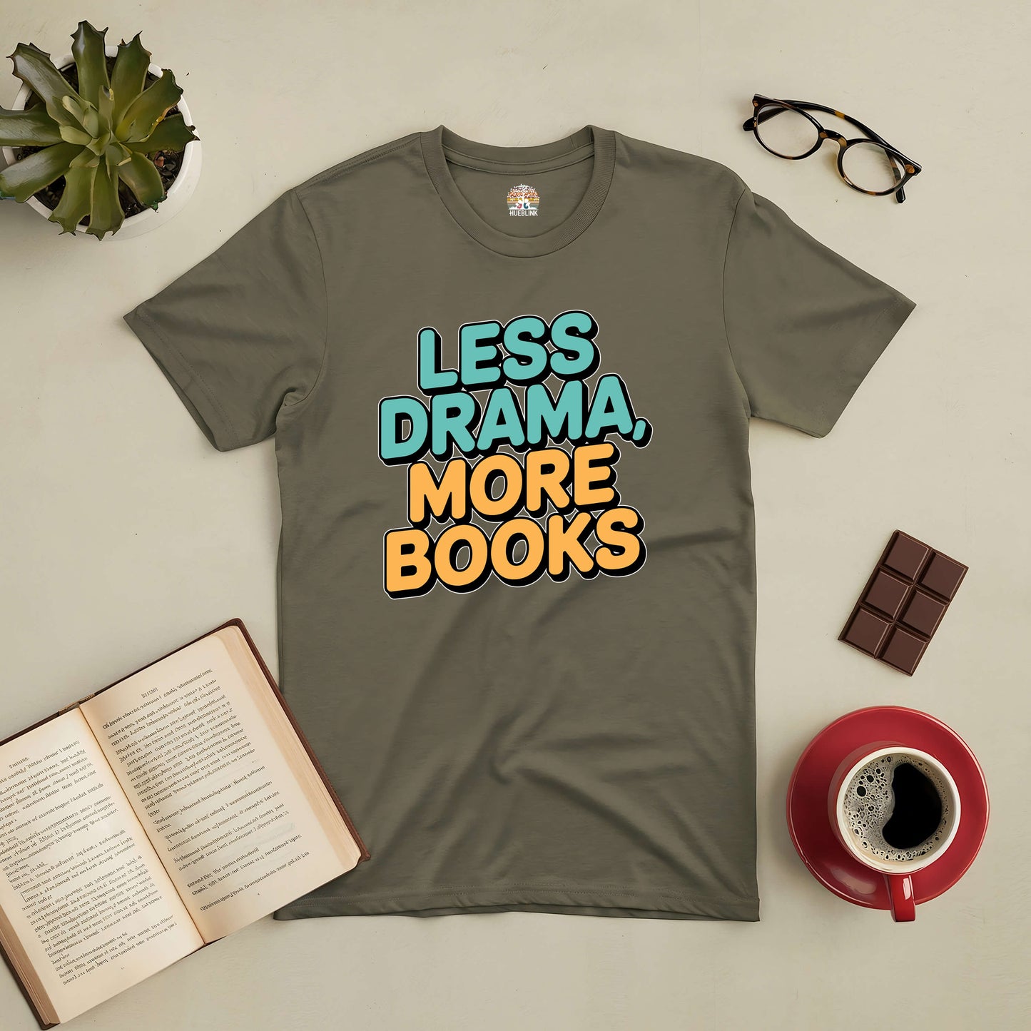 Less Drama More Books tee laid flat with open book, glasses, coffee cup, and chocolate bar on a desk.