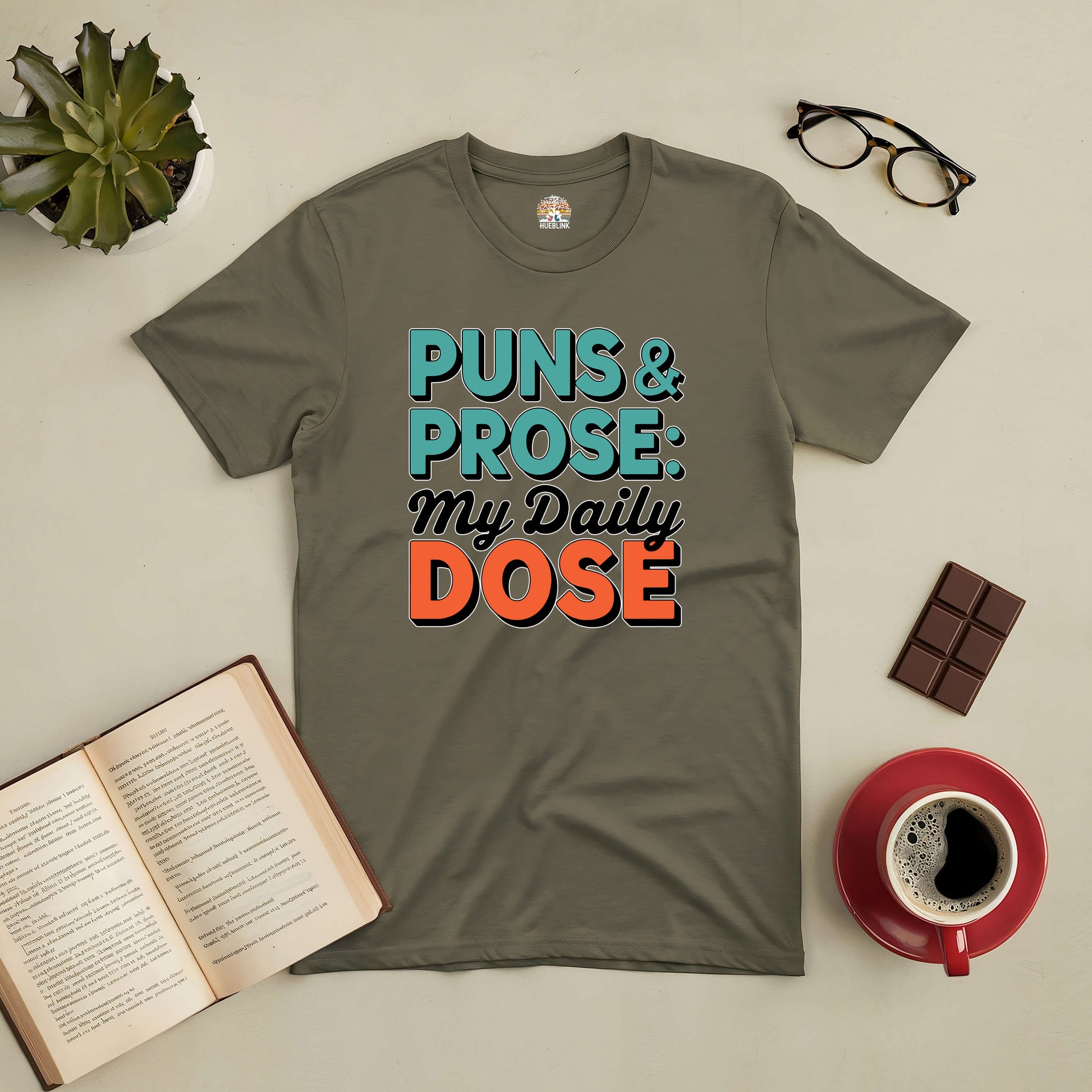 "Puns & Prose My Daily Dose Tee with witty wordplay design, surrounded by coffee, a book, glasses, and chocolate."