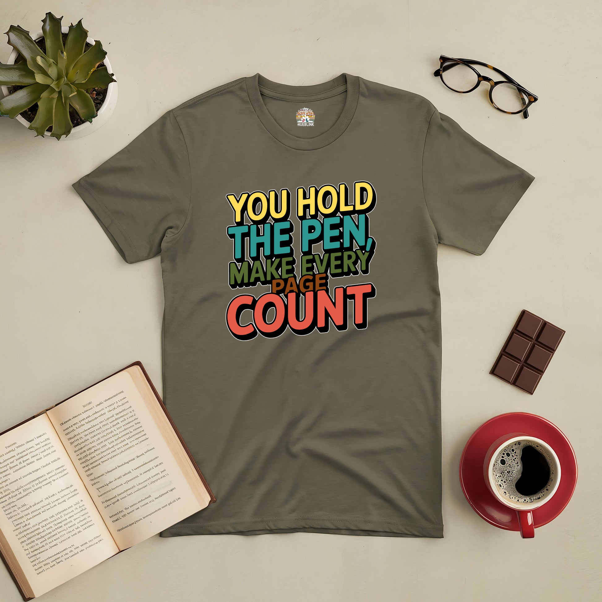 Inspirational "You Hold the Pen, Make Every Page Count" tee on table with open book, coffee, glasses, plant, and chocolate.