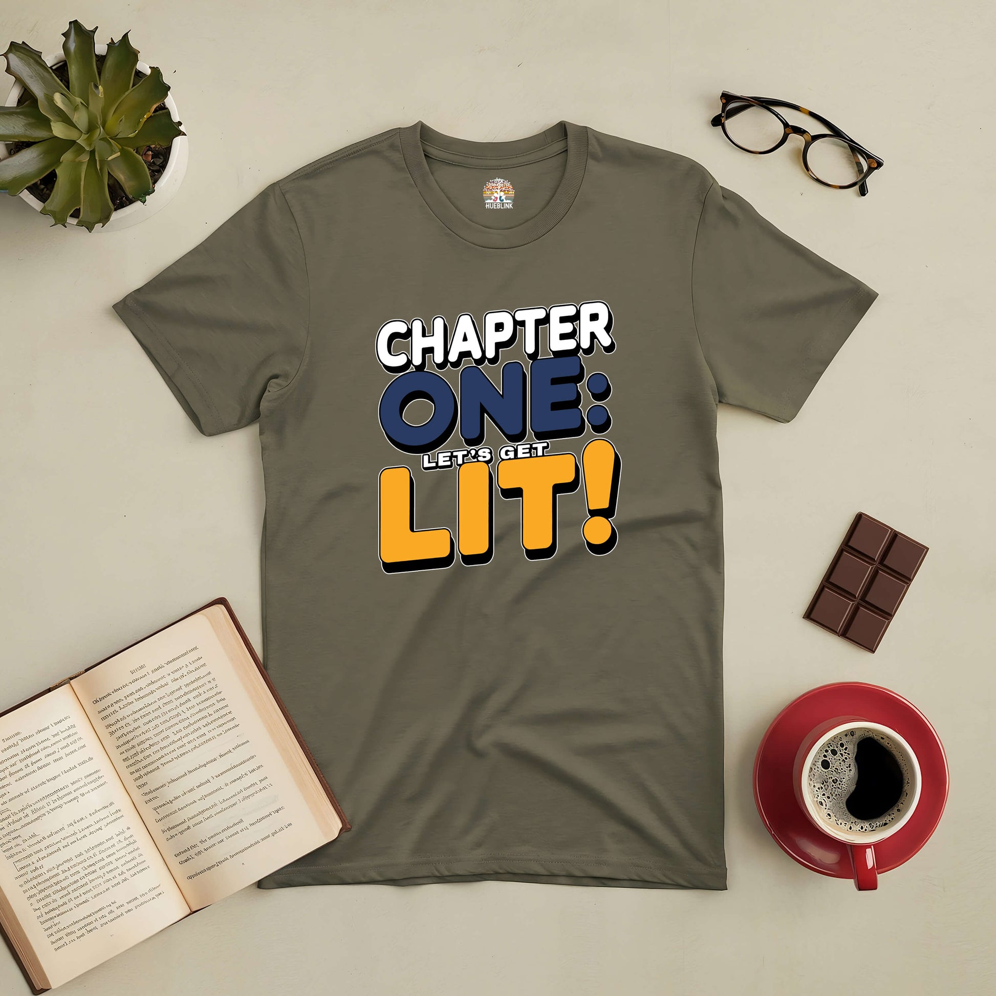 Olive green T-shirt with "Chapter One: Let's Get Lit!" text, surrounded by a book, coffee, chocolate, glasses, and a plant.