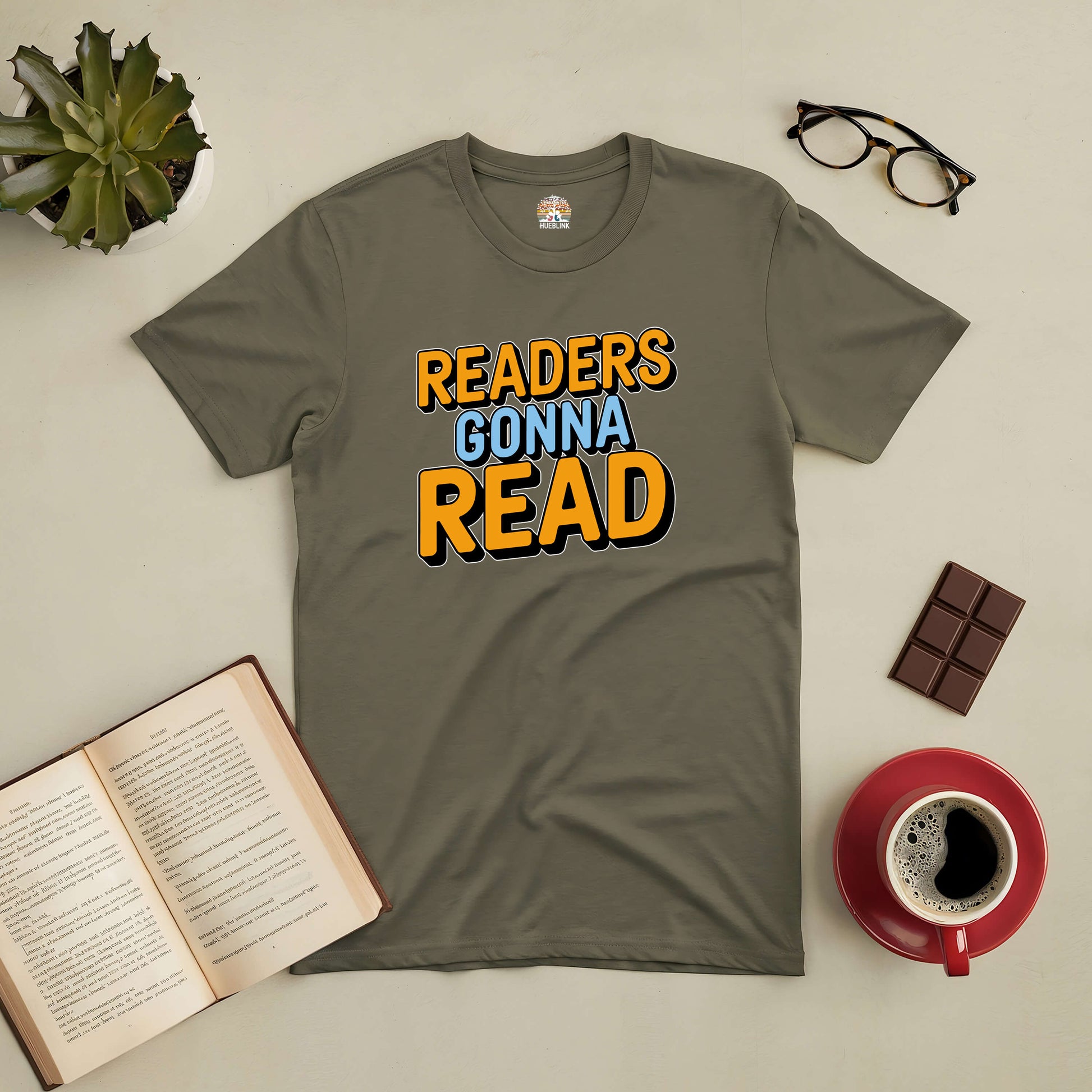 "Readers Gonna Read Tee with playful typography, surrounded by book, glasses, chocolate, and coffee on a table"