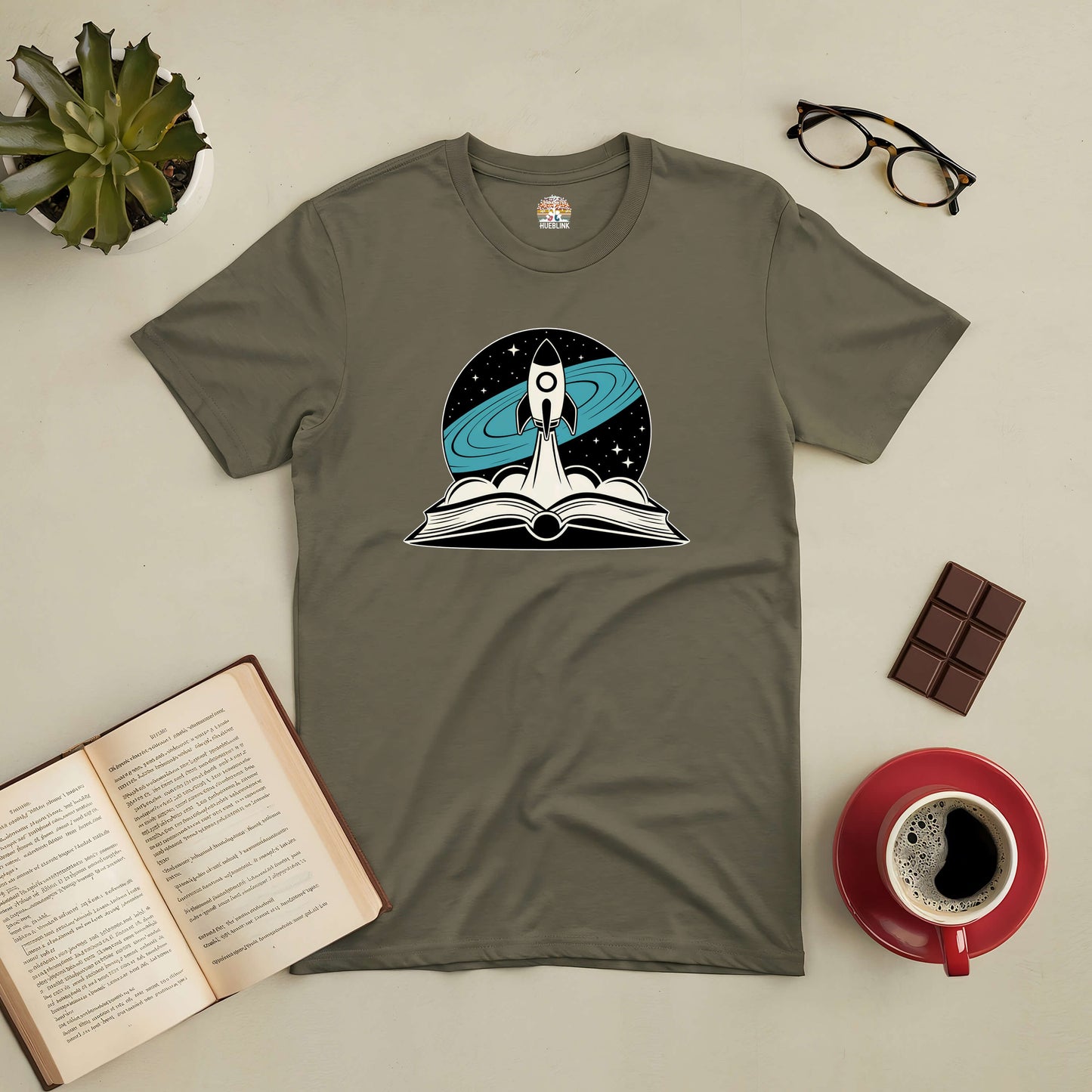 Rocket Science tee with rocket launching from book, surrounded by coffee, glasses, and chocolate on a desk.