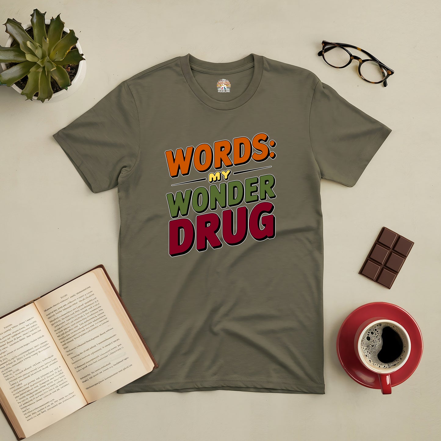 Olive green tee with "Words: My Wonder Drug" text, surrounded by book, coffee, glasses, and chocolate on a flat surface.