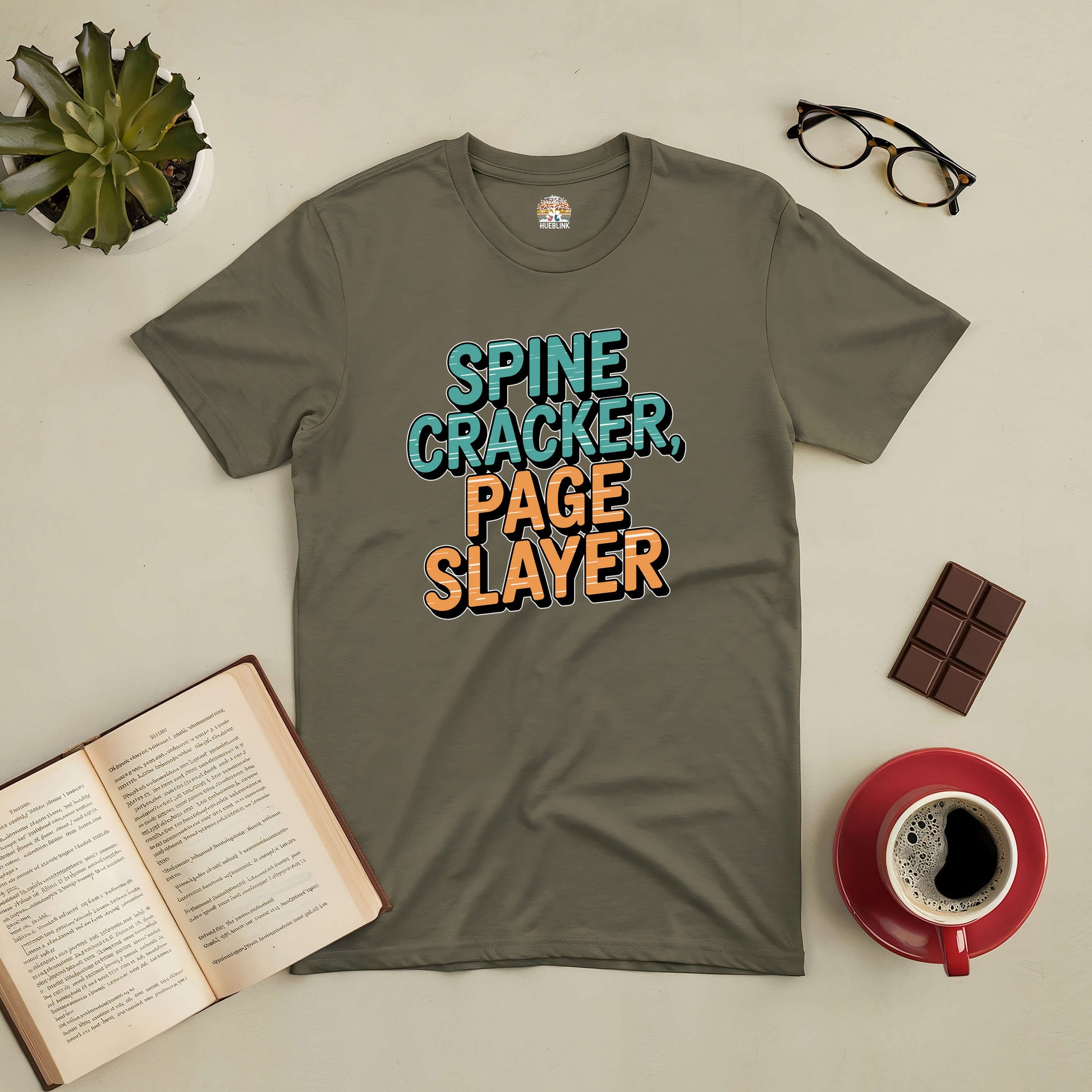 Spine Cracker, Page Slayer tee for book lovers with open book, glasses, chocolate, coffee, and plant on a table.