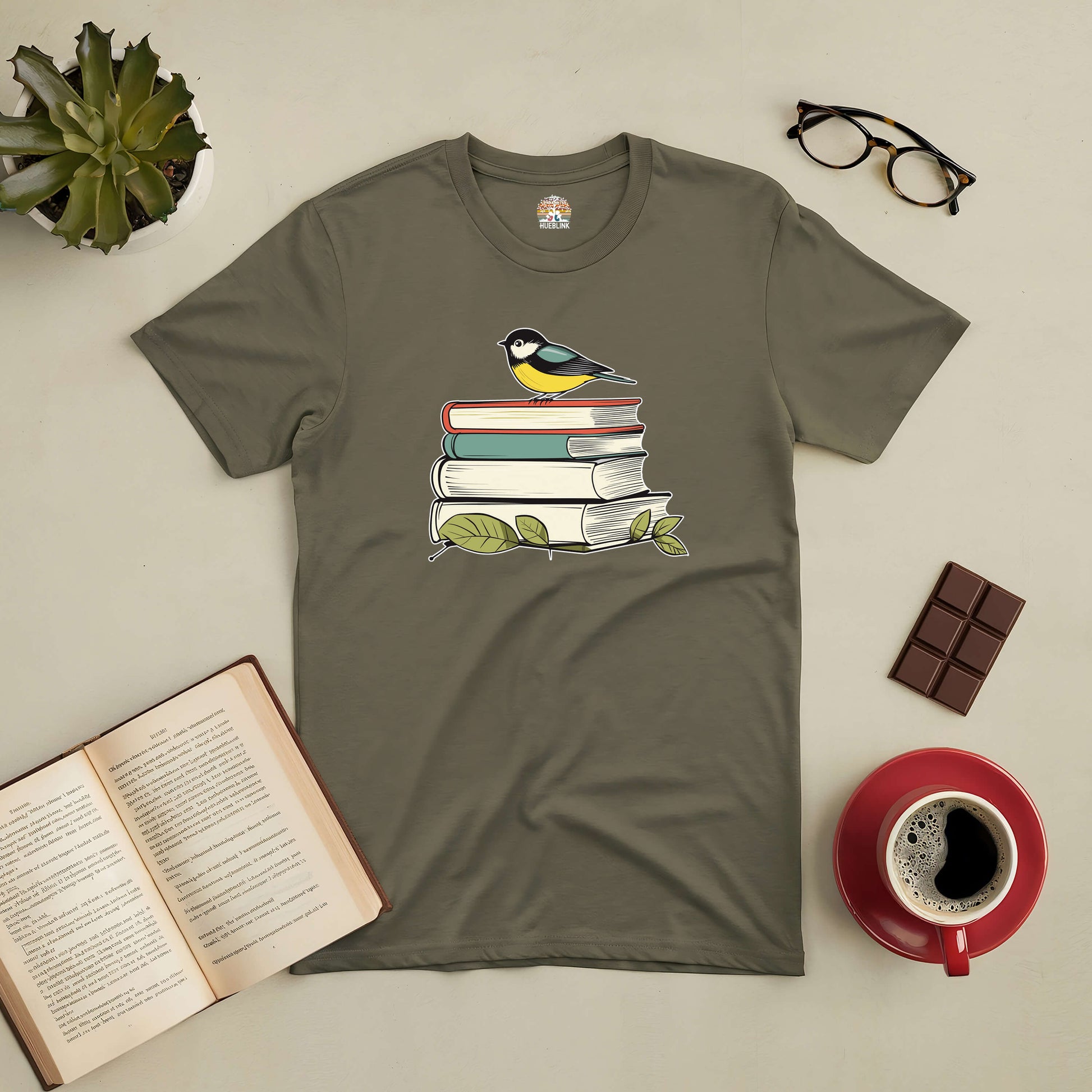 Olive green tee with bird on books graphic, surrounded by open book, coffee, chocolate, glasses, and plant. Perfect for book lovers.