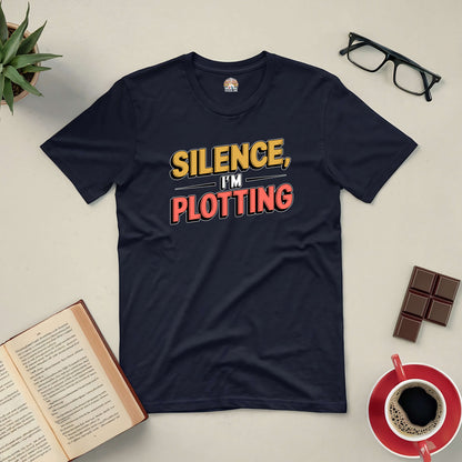 Black t-shirt with bold "Silence, I'm Plotting" text, surrounded by glasses, open book, chocolate, coffee, and plant.