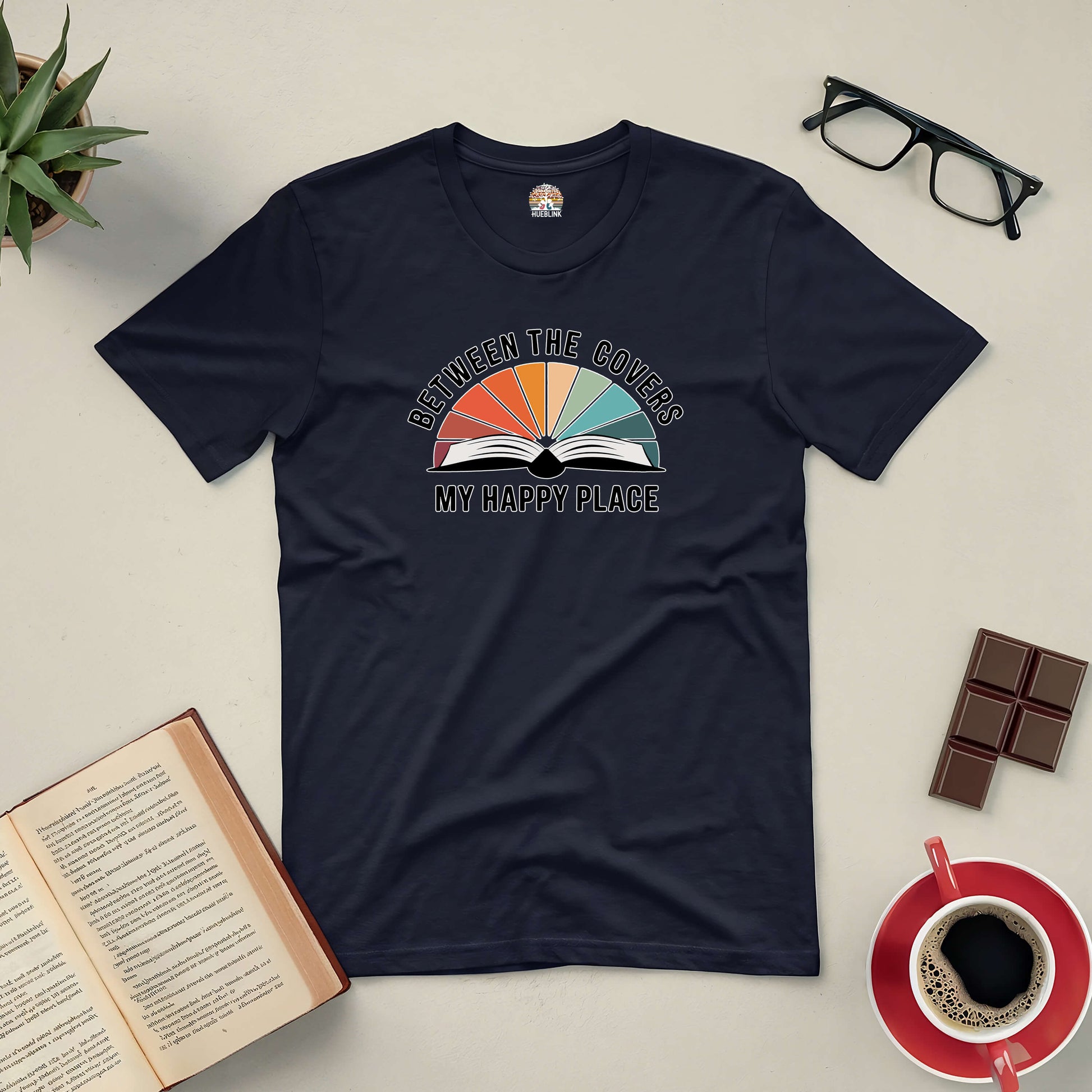 Black "Between the Covers: My Happy Place" tee with an open book graphic, surrounded by glasses, chocolate, coffee, and a potted plant.