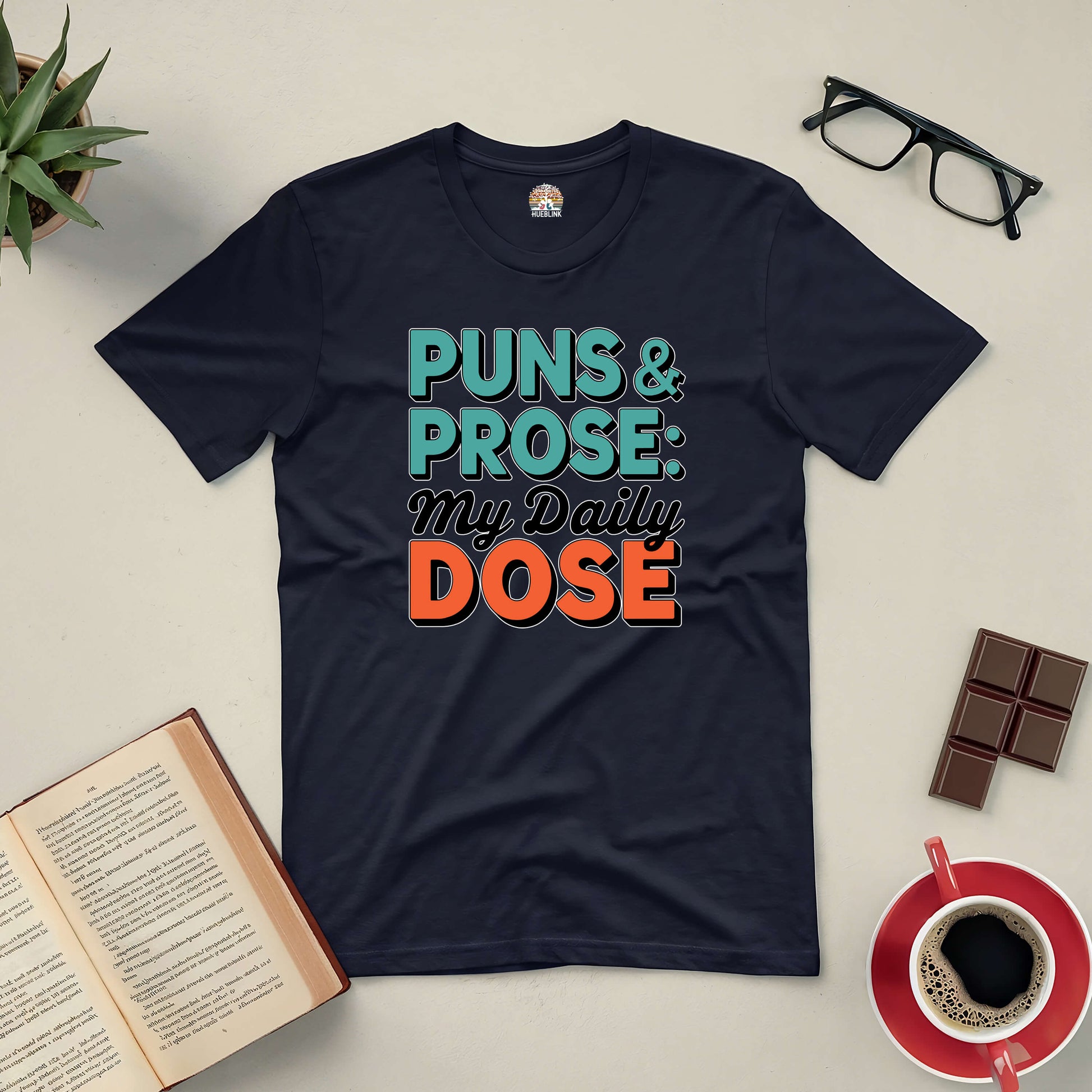 Black tee with "Puns & Prose: My Daily Dose" text, surrounded by book, glasses, coffee, and chocolate on a light background.