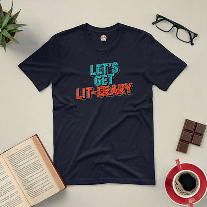 Black "Let's Get Lit-erary" tee with colorful text, surrounded by open book, glasses, chocolate, coffee, and plant on a beige background.