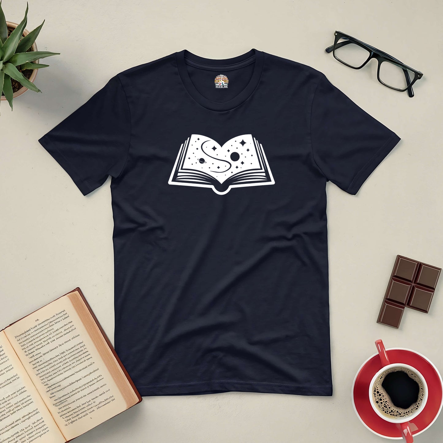 Black t-shirt featuring a cosmic book design, surrounded by coffee, chocolate, glasses, and open books, celebrating imagination.